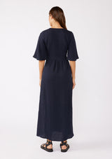 [Color: Navy] A brunette model wearing a textured maxi dress in a deep navy blue. With a deep v neckline, empire waistline, flowy half sleeves, and a side slit. Paired with casual sandals for any daytime outing.