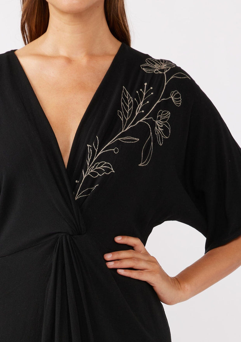 [Color: Black/Natural] A brunette model wearing a black front knot maxi dress with a contrast embroidery, Featuring a deep v neckline, kimono sleeves, front slit, and an asymmetrical hem. A bohemian style maxi dress with a relaxed fit.