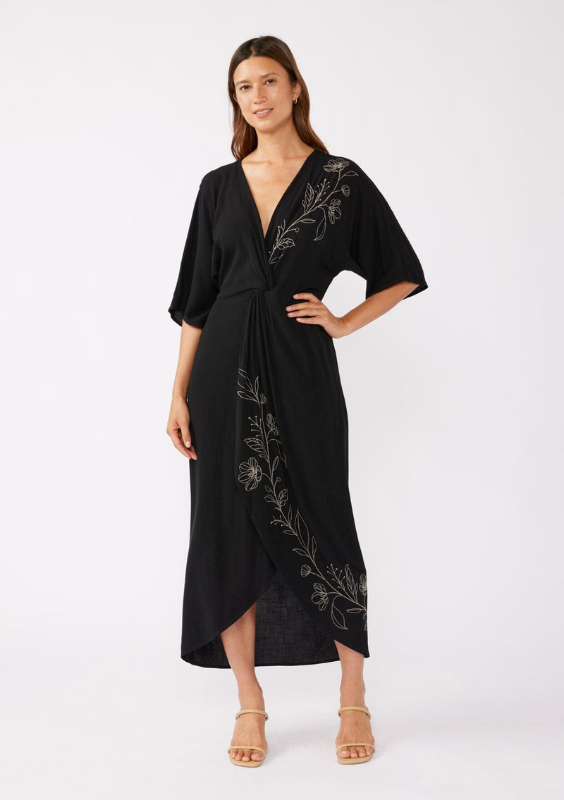 [Color: Black/Natural] A brunette model wearing a black front knot maxi dress with a contrast embroidery, Featuring a deep v neckline, kimono sleeves, front slit, and an asymmetrical hem. A bohemian style maxi dress with a relaxed fit.  