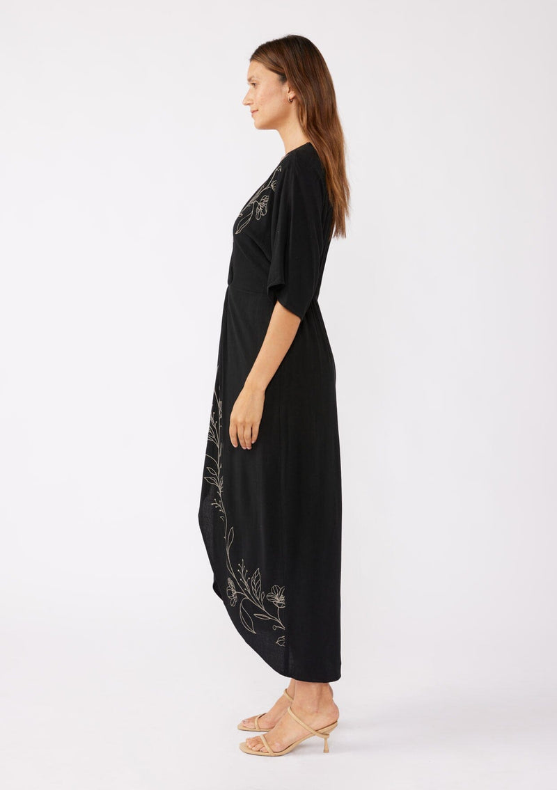 [Color: Black/Natural] A brunette model wearing a black front knot maxi dress with a contrast embroidery, Featuring a deep v neckline, kimono sleeves, front slit, and an asymmetrical hem. A bohemian style maxi dress with a relaxed fit.