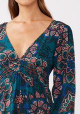 [Color: Teal/Rust] A brunette model wearing a teal floral mini dress with an open back and tie detail. An empire waist mini dress with long sleeves, a deep v neckline, and smocked cuffs. The perfect bohemian mini dress for the Fall season. 