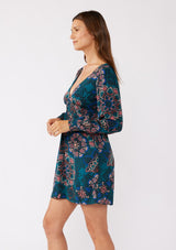 [Color: Teal/Rust] A brunette model wearing a teal floral mini dress with an open back and tie detail. An empire waist mini dress with long sleeves, a deep v neckline, and smocked cuffs. The perfect bohemian mini dress for the Fall season. 