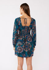 [Color: Teal/Rust] A brunette model wearing a teal floral mini dress with an open back and tie detail. An empire waist mini dress with long sleeves, a deep v neckline, and smocked cuffs. The perfect bohemian mini dress for the Fall season. 