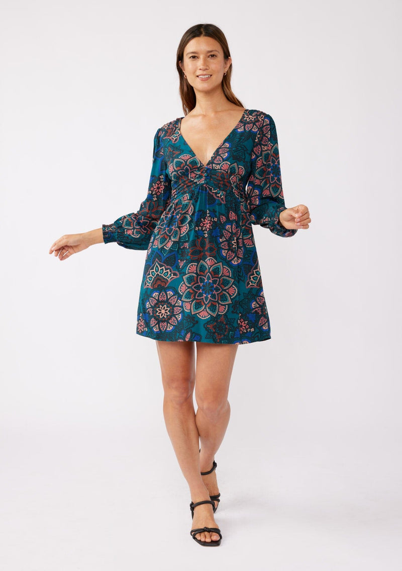 [Color: Teal/Rust] A brunette model wearing a teal floral mini dress with an open back and tie detail. An empire waist mini dress with long sleeves, a deep v neckline, and smocked cuffs. The perfect bohemian mini dress for the Fall season. 