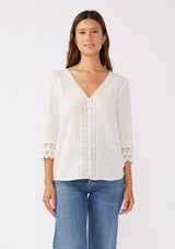 [Color: Vanilla] A blonde model wearing a textured cotton blouse in ivory white. With lace and crochet trims, functional button front, v neckline, and relaxed fit. A casual bohemian top paired with denim jeans.