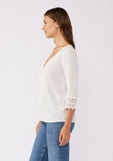 [Color: Vanilla] A blonde model wearing a textured cotton blouse in ivory white. With lace and crochet trims, functional button front, v neckline, and relaxed fit. A casual bohemian top paired with denim jeans.