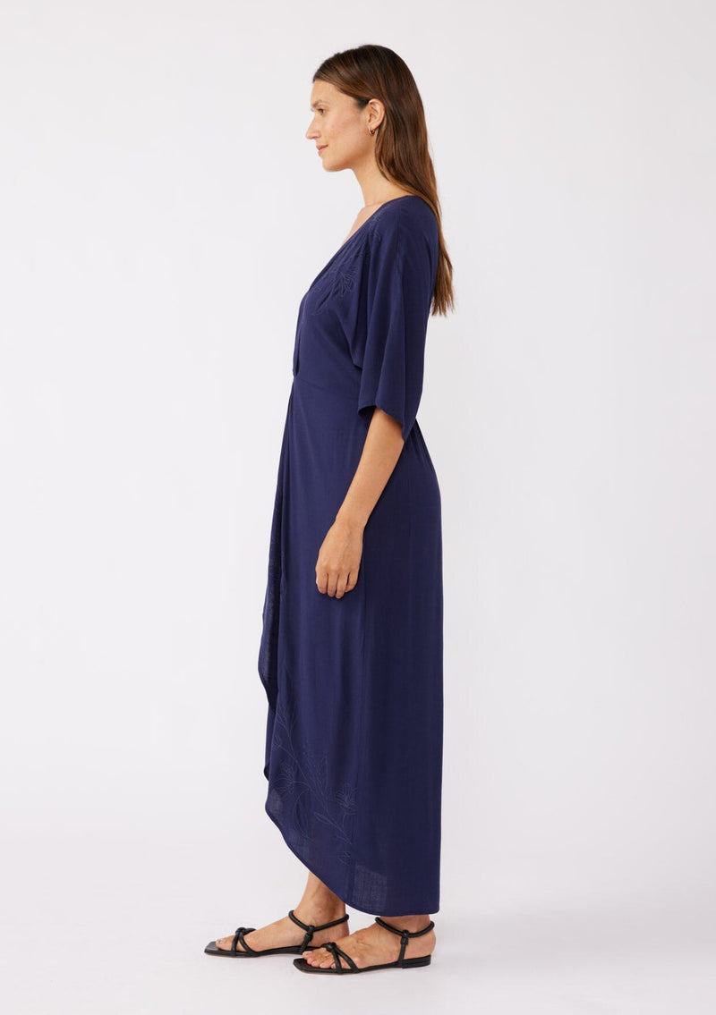 [Color: Navy/Navy] A brunette model wearing a blue front knot maxi dress with a tonal embroidery. Featuring a deep v neckline, kimono sleeves, front slit, and an asymmetrical hem. A bohemian style maxi dress with a relaxed fit.