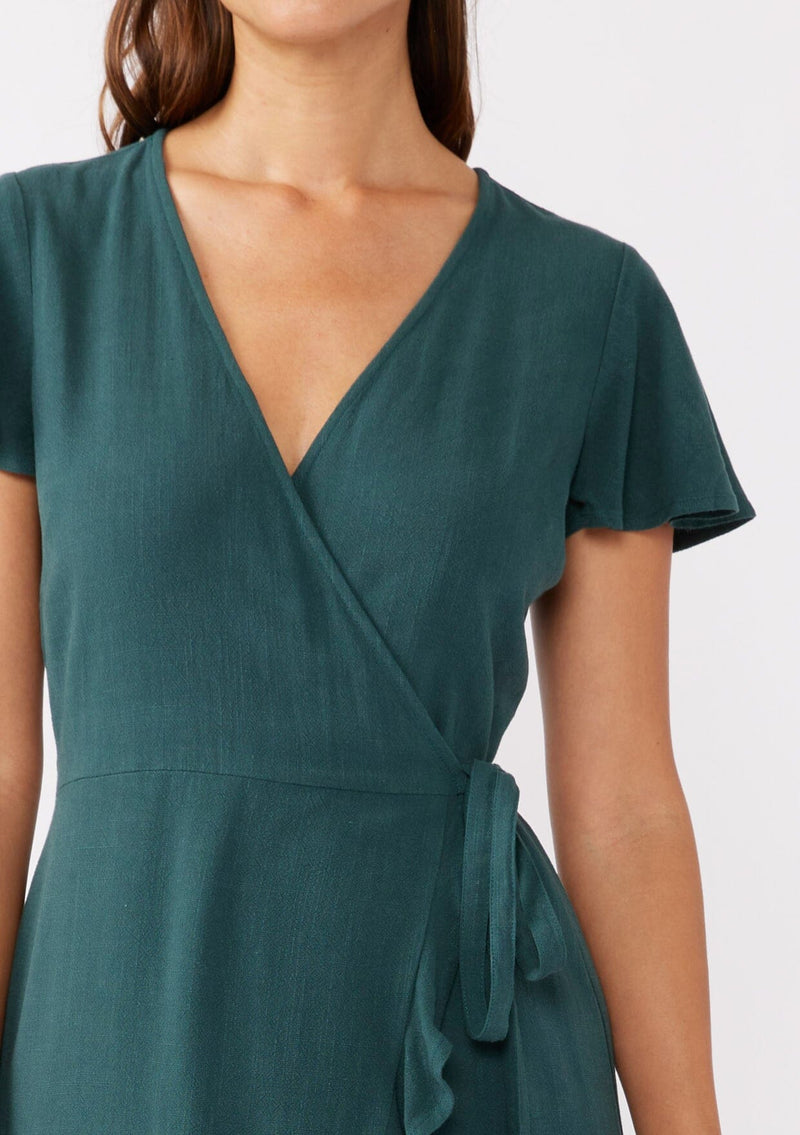 [Color: Dusty Teal] A brunette model wearing a classic short sleeve wrap dress in a solid autumn green hue. With a surplice v neckline, side tie wrap closure, and a ruffle trimmed hem. A rayon linen blend mini dress that's versatile and can be worn to many special occasions.