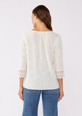 [Color: Vanilla] A blonde model wearing a textured cotton blouse in ivory white. With lace and crochet trims, functional button front, v neckline, and relaxed fit. A casual bohemian top paired with denim jeans.