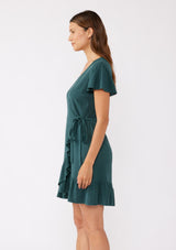 [Color: Dusty Teal] A brunette model wearing a classic short sleeve wrap dress in a solid autumn green hue. With a surplice v neckline, side tie wrap closure, and a ruffle trimmed hem. A rayon linen blend mini dress that's versatile and can be worn to many special occasions.