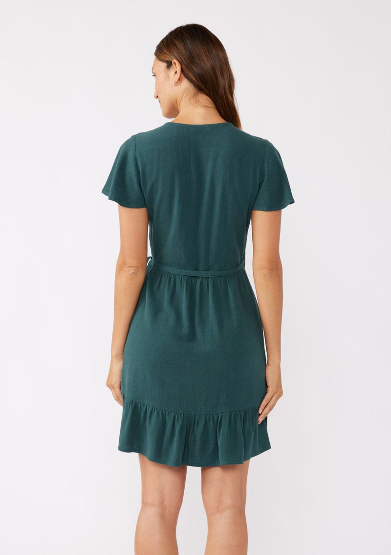 [Color: Dusty Teal] A brunette model wearing a classic short sleeve wrap dress in a solid autumn green hue. With a surplice v neckline, side tie wrap closure, and a ruffle trimmed hem. A rayon linen blend mini dress that's versatile and can be worn to many special occasions.