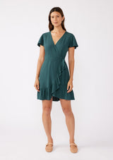 [Color: Dusty Teal] A brunette model wearing a classic short sleeve wrap dress in a solid autumn green hue. With a surplice v neckline, side tie wrap closure, and a ruffle trimmed hem. A rayon linen blend mini dress that's versatile and can be worn to many special occasions.