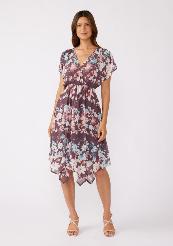 [Color: Plum/Blue] A brunette model wearing an ultra-flowy bohemian midi dress with an all over floral print. A printed deep dark purple midi dress with short dolman sleeves, v neckline, open back with tie detail, and an asymmetrical hemline. Perfect for special occasions and events during the fall season. 