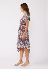 [Color: Plum/Blue] A brunette model wearing an ultra-flowy bohemian midi dress with an all over floral print. A printed deep dark purple midi dress with short dolman sleeves, v neckline, open back with tie detail, and an asymmetrical hemline. Perfect for special occasions and events during the fall season. 