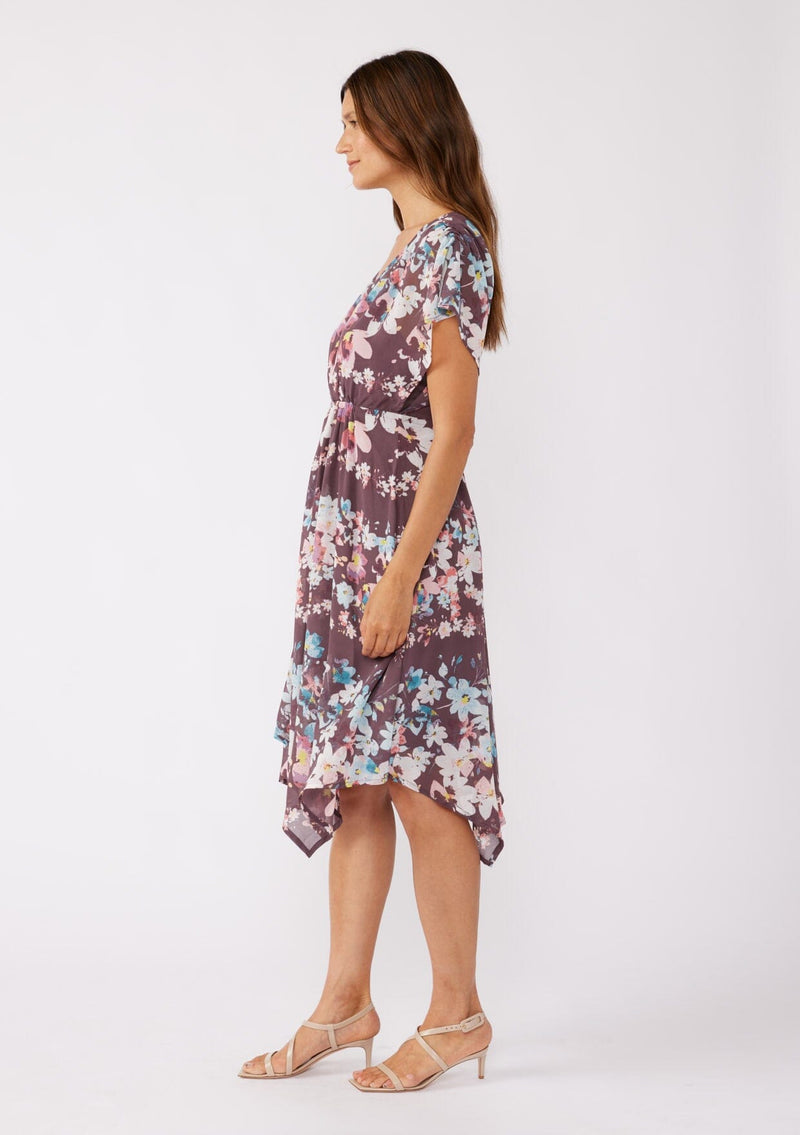 [Color: Plum/Blue] A brunette model wearing an ultra-flowy bohemian midi dress with an all over floral print. A printed deep dark purple midi dress with short dolman sleeves, v neckline, open back with tie detail, and an asymmetrical hemline. Perfect for special occasions and events during the fall season. 