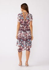 [Color: Plum/Blue] A brunette model wearing an ultra-flowy bohemian midi dress with an all over floral print. A printed deep dark purple midi dress with short dolman sleeves, v neckline, open back with tie detail, and an asymmetrical hemline. Perfect for special occasions and events during the fall season. 