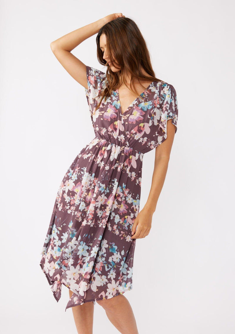 [Color: Plum/Blue] A brunette model wearing an ultra-flowy bohemian midi dress with an all over floral print. A printed deep dark purple midi dress with short dolman sleeves, v neckline, open back with tie detail, and an asymmetrical hemline. Perfect for special occasions and events during the fall season. 