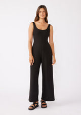 [Color: Black] A brunette model wearing an all black jumpsuit with a slim fit bodice and wide legs. Featuring a flattering scoop neckline and back and thick tank top straps. Paired casually with thick strappy sandals for a casual look. An ideal jumpsuit for sophisticated events or office. 