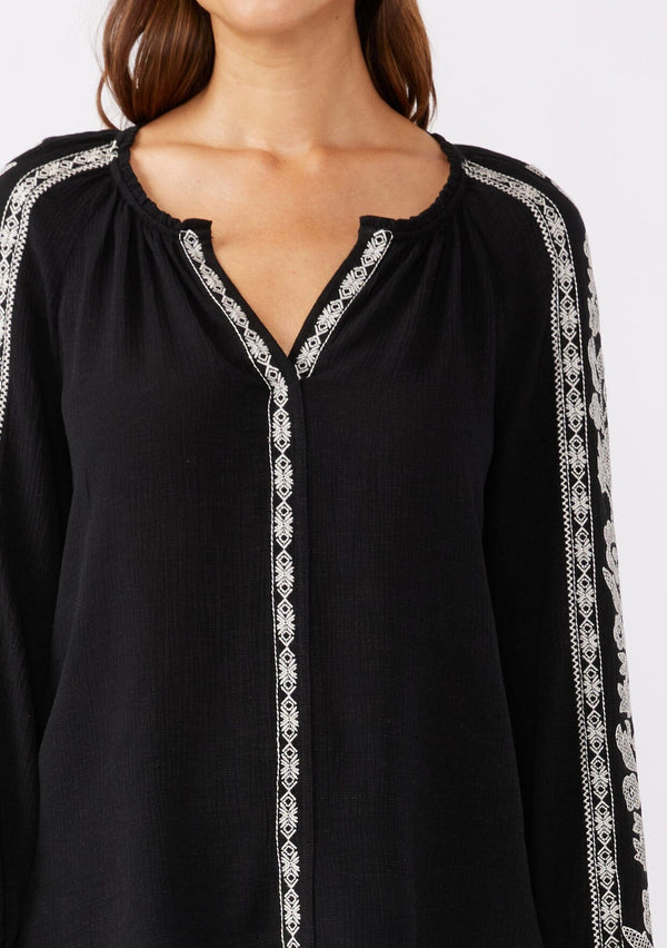 [Color: Black/Natural] A brunette model wearing an ultra bohemian blouse in black with off white embroidery. Featuring a split v neckline, long sleeves, and a relaxed fit.
