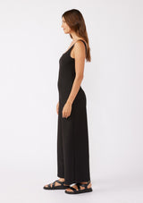 [Color: Black] A brunette model wearing an all black jumpsuit with a slim fit bodice and wide legs. Featuring a flattering scoop neckline and back and thick tank top straps. Paired casually with thick strappy sandals for a casual look. An ideal jumpsuit for sophisticated events or office.