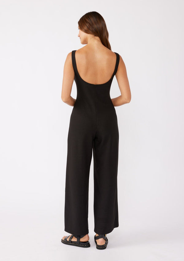 [Color: Black] A brunette model wearing an all black jumpsuit with a slim fit bodice and wide legs. Featuring a flattering scoop neckline and back and thick tank top straps. Paired casually with thick strappy sandals for a casual look. An ideal jumpsuit for sophisticated events or office.