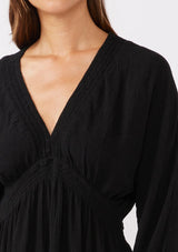 [Color: Black] A detail image of a brunette model wearing an ultra flowy black mini dress with a relaxed silhouette. With a v neckline with a smocked edge detail running through the waist, long dolman sleeves, and convenient side pockets. 