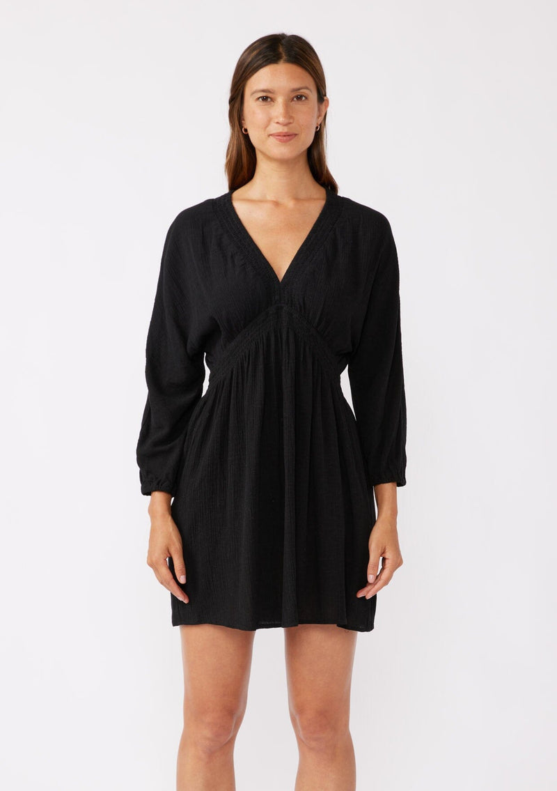 [Color: Black] A back image of a brunette model wearing an ultra flowy black mini dress with a relaxed silhouette. With a v neckline with a smocked edge detail running through the waist, long dolman sleeves, and convenient side pockets.