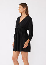 [Color: Black] A side image of a brunette model wearing an ultra flowy black mini dress with a relaxed silhouette. With a v neckline with a smocked edge detail running through the waist, long dolman sleeves, and convenient side pockets.