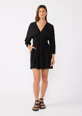 [Color: Black] A brunette model wearing an ultra flowy black mini dress with a relaxed silhouette. With a v neckline with a smocked edge detail running through the waist, long dolman sleeves, and convenient side pockets. Paired with trendy thick strappy sandals for an elevated casual look.  