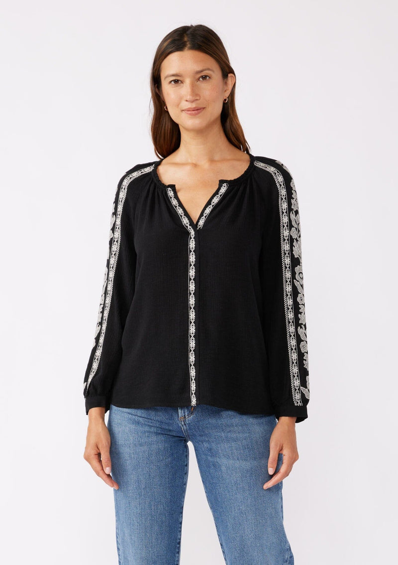 [Color: Black/Natural] A brunette model wearing an ultra bohemian blouse in black with off white embroidery. Featuring a split v neckline, long sleeves, and a relaxed fit.