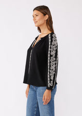 [Color: Black/Natural] A brunette model wearing an ultra bohemian blouse in black with off white embroidery. Featuring a split v neckline, long sleeves, and a relaxed fit.