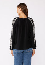 [Color: Black/Natural] A brunette model wearing an ultra bohemian blouse in black with off white embroidery. Featuring a split v neckline, long sleeves, and a relaxed fit.