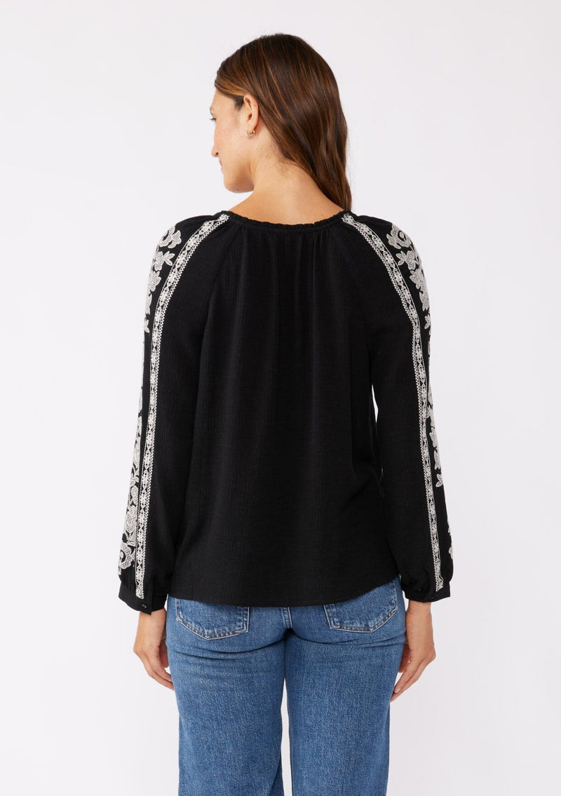 [Color: Black/Natural] A brunette model wearing an ultra bohemian blouse in black with off white embroidery. Featuring a split v neckline, long sleeves, and a relaxed fit.