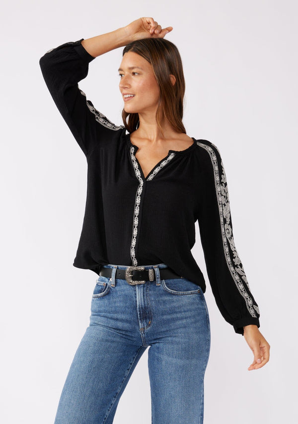 [Color: Black/Natural] A brunette model wearing an ultra bohemian blouse in black with off white embroidery. Featuring a split v neckline, long sleeves, and a relaxed fit. 