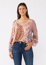 [Color: Natural/Clay] A brunette model wearing a bohemian long sleeve blouse with a pink patchwork print. A relaxed fit style with a classic v neckline, elastic cuffs, pintuck details, and unique burlap trim inserts. Perfectly styled with dark denim and tucked in for a laid back vibe. 