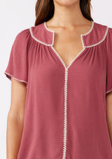 [Color: Mauve/Natural] A brunette model wearing a classic bohemian top in a mauve pink hue. With an embroidered blanket stitch trim, short flutter sleeves, and a split v neckline. Perfect for any fall wardrobe; styled with denim for a cool and casual look.  