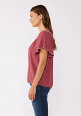 [Color: Mauve/Natural] A brunette model wearing a classic bohemian top in a mauve pink hue. With an embroidered blanket stitch trim, short flutter sleeves, and a split v neckline. Perfect for any fall wardrobe; styled with denim for a cool and casual look.  