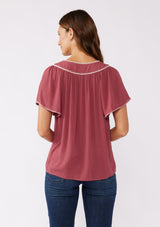 [Color: Mauve/Natural] A brunette model wearing a classic bohemian top in a mauve pink hue. With an embroidered blanket stitch trim, short flutter sleeves, and a split v neckline. Perfect for any fall wardrobe; styled with denim for a cool and casual look.  