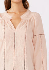 [Color: Dusty Rose] A detailed image of a brunette model wearing a pretty boho blouse in a dusty pink. With floral tonal embroidery, lace trim inserts, long sleeves with smocked wrist cuffs, and a split v neckline. An airy and relaxed fit blouse crafted in a cotton blend fabric. 