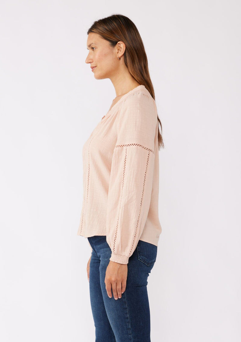 [Color: Dusty Rose] A side facing image of a brunette model wearing a pretty boho blouse in a dusty pink. With floral tonal embroidery, lace trim inserts, long sleeves with smocked wrist cuffs, and a split v neckline. An airy and relaxed fit blouse crafted in a cotton blend fabric. 