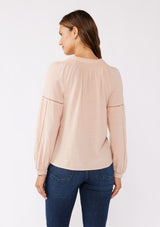 [Color: Dusty Rose] A back facing image of a brunette model wearing a pretty boho blouse in a dusty pink. With floral tonal embroidery, lace trim inserts, long sleeves with smocked wrist cuffs, and a split v neckline. An airy and relaxed fit blouse crafted in a cotton blend fabric. 