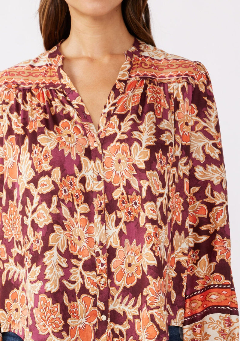 [Color: Wine/Dusty Rose] A detailed image of a brunette model wearing an orange and red floral print blouse with a functional button front. With a relaxed fit, split v neckline, long sleeves, and elastic cuffs. 