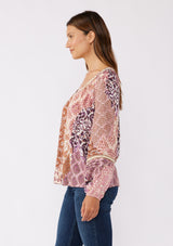 [Color: Natural/Clay] A side image of brunette model wearing a bohemian long sleeve blouse with a pink patchwork print. A relaxed fit style with a classic v neckline, elastic cuffs, pintuck details, and unique burlap trim inserts. Perfectly styled with dark denim for a laid back fall vibe. 