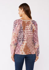 [Color: Natural/Clay] A back image of brunette model wearing a bohemian long sleeve blouse with a pink patchwork print. A relaxed fit style with a classic v neckline, elastic cuffs, pintuck details, and unique burlap trim inserts. Perfectly styled with dark denim for a laid back fall vibe. 