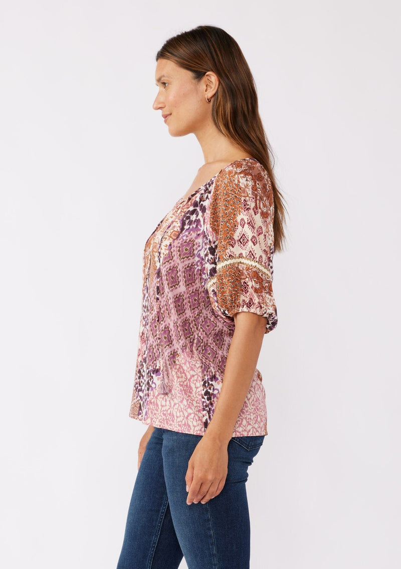 [Color: Natural/Clay] A brunette model wearing a bohemian puff sleeve top in a bohemian pink patchwork print. Featuring short puff sleeves, split v neckline with ties, and burlap trim inserts at the sleeve. A classic boho top styled with denim for the fall season. 