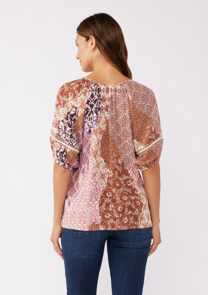 [Color: Natural/Clay] A brunette model wearing a bohemian puff sleeve top in a bohemian pink patchwork print. Featuring short puff sleeves, split v neckline with ties, and burlap trim inserts at the sleeve. A classic boho top styled with denim for the fall season. 