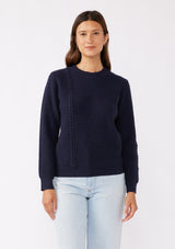 [Color: Navy] A brunette model wearing a classic crew neck sweater in navy blue. With ribbed trims, long sleeves, and a cable knit stitch detail. A versatile sweater perfect for layering during colder temperatures or wearing on its own. 