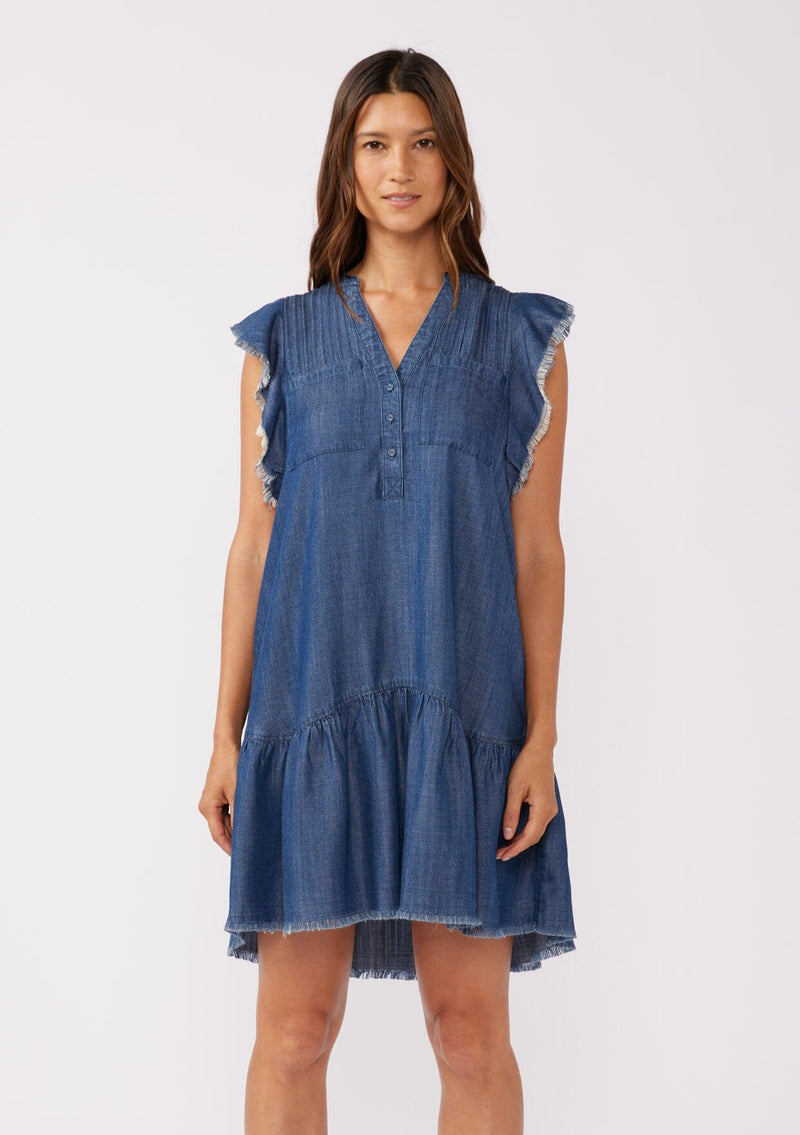 [Color: Midnight Wash] A front facing image of a brunette model wearing a denim blue bohemian fall mini dress crafted from Tencel. With short flutter sleeves, a v neckline, a tiered mini skirt, front patch pockets, a self covered button front, a raw hemline, and pleated details.