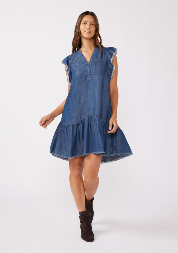 [Color: Midnight Wash] A front facing image of a brunette model wearing a denim blue bohemian fall mini dress crafted from Tencel. With short flutter sleeves, a v neckline, a tiered mini skirt, front patch pockets, a self covered button front, a raw hemline, and pleated details.