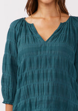 [Color: Deep Teal] A detailed image of a brunette model wearing a textured jacquard stripe blouse in teal. A relaxed fit blouse with a split v neckline and long sleeves. Styled with dark denim for the fall season. 